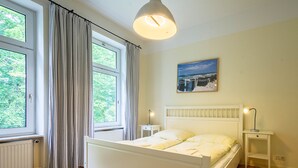 Ein Schlafzimmer mit Bett, Nachttisch, Vorhängen am Fenster, Deckenleuchte und einem Bild an der Wand.