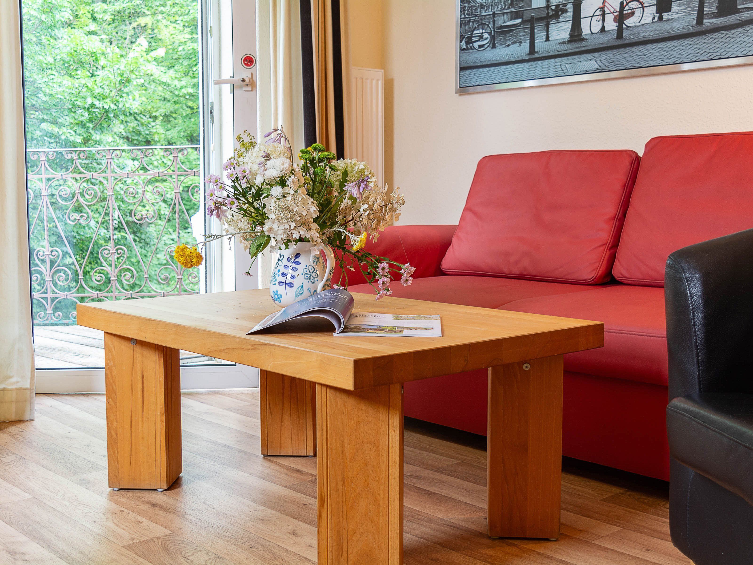 A wooden table with a vase of flowers, a magazine, and a red sofa in a room with wooden flooring and a view of greenery through a window.