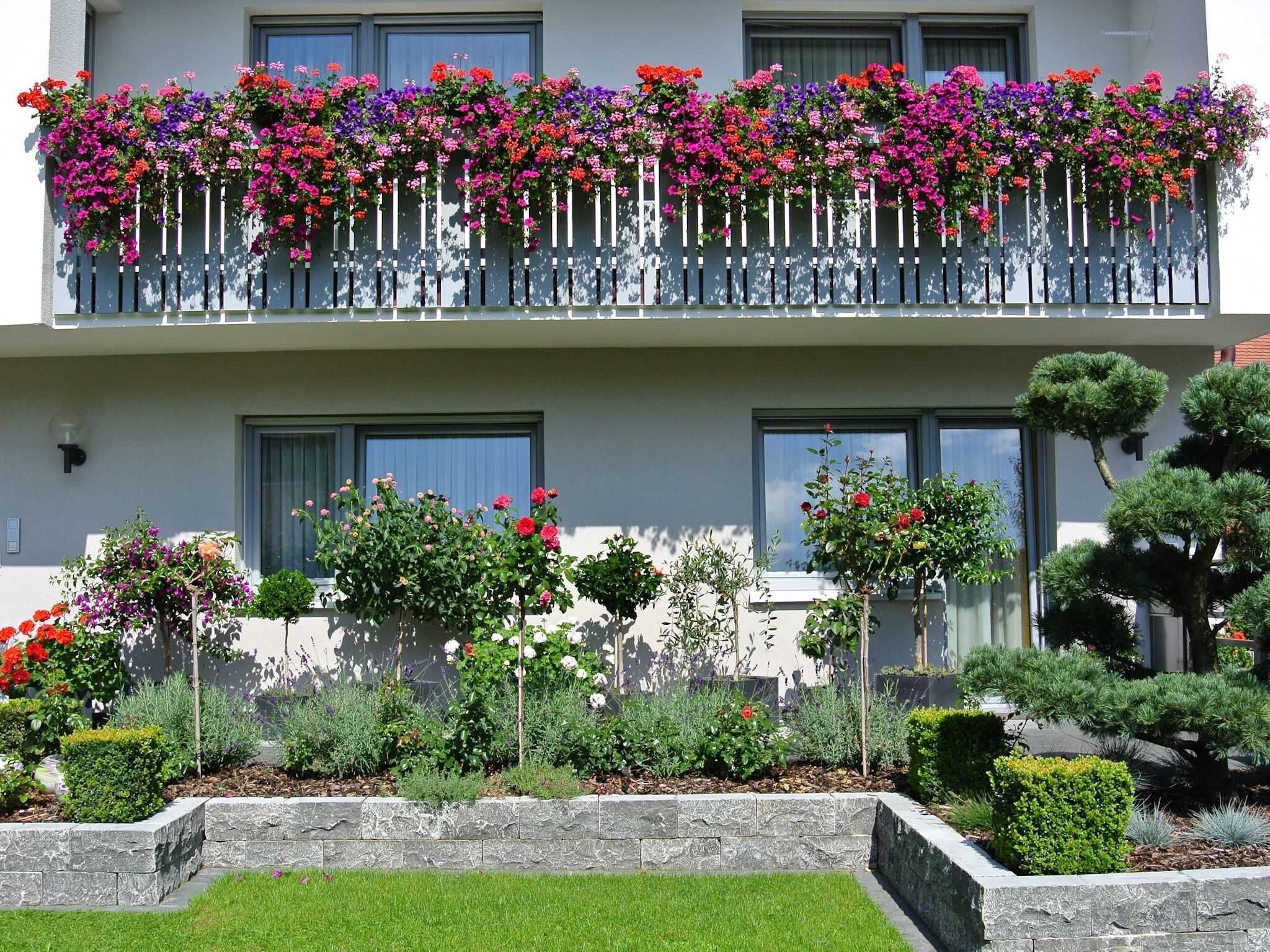 Ein zweistöckiges Gebäude mit einem Balkon, der mit bunten Blumen geschmückt ist, ein gepflegter Garten mit verschiedenen Pflanzen und eine Steinmauer.