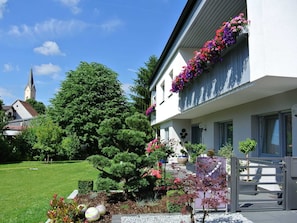 Un bâtiment blanc doté d’un balcon orné de fleurs colorées, un jardin bien entretenu avec différentes espèces de plantes et, au loin, le clocher d’une église.