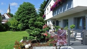 A white building with a balcony adorned with colorful flowers, a well-maintained garden with various plants, and a church steeple in the background.