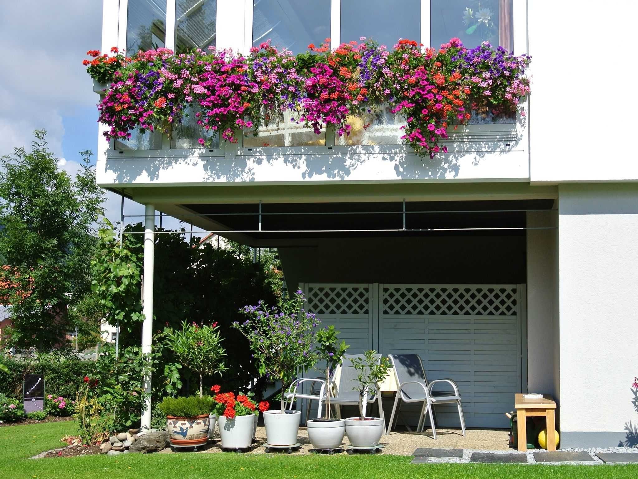 A two-story building with a balcony filled with colorful flowers, a garage door, and potted plants in the front yard.