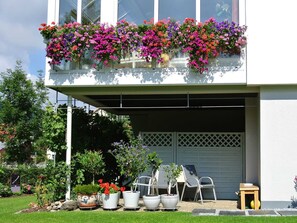 A two-story building with a balcony filled with colorful flowers, a garage door, and potted plants in the front yard. - Apartment on the Wiesent - Apartment An der Wiesent (Ebermannstadt)