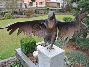 A bird sculpture on a pedestal in a garden with a hedge and a building in the background.