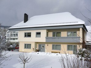 A two-story building with a snow-covered roof, a balcony on the second floor, and a front door with a wreath. - Apartment on the Wiesent - Apartment An der Wiesent (Ebermannstadt)