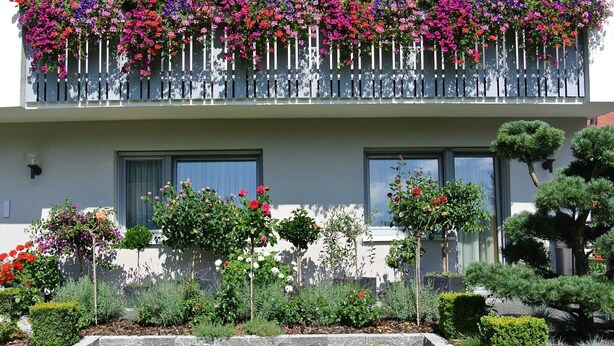 A two-story building with a balcony adorned with colorful flowers, a well-maintained garden with various plants, and a stone retaining wall.