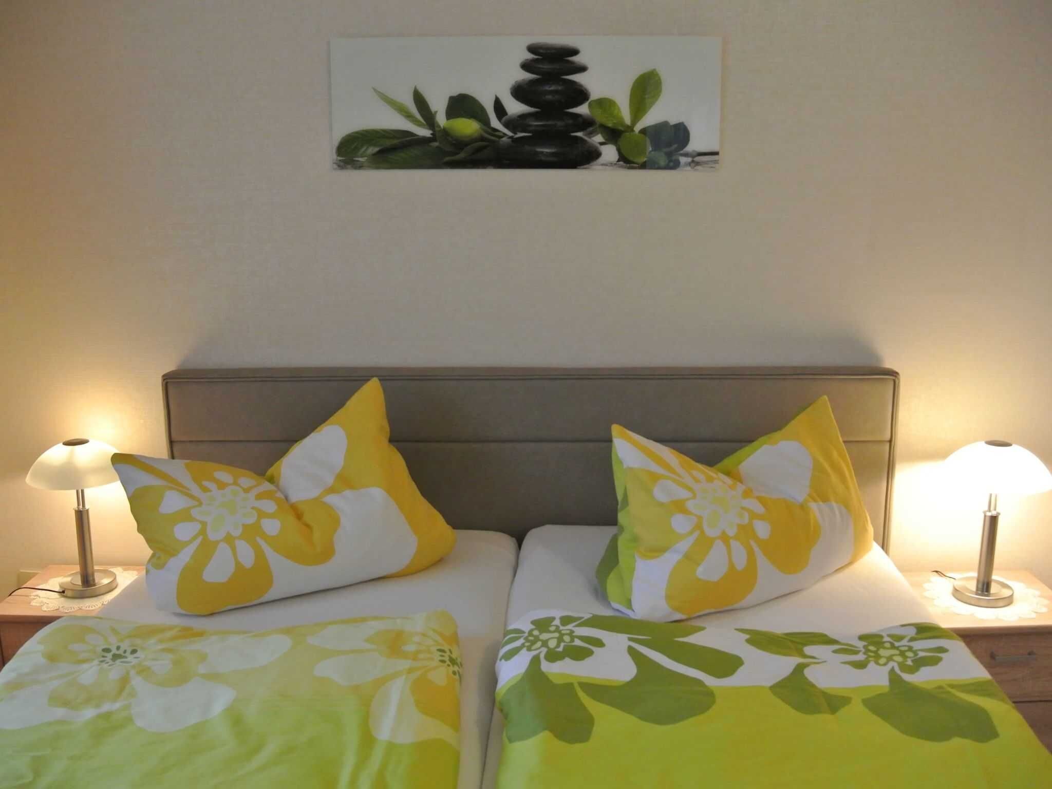 A double bed with floral bedding, two pillows with a yellow and white pattern, and a wall-mounted picture of stones and plants.