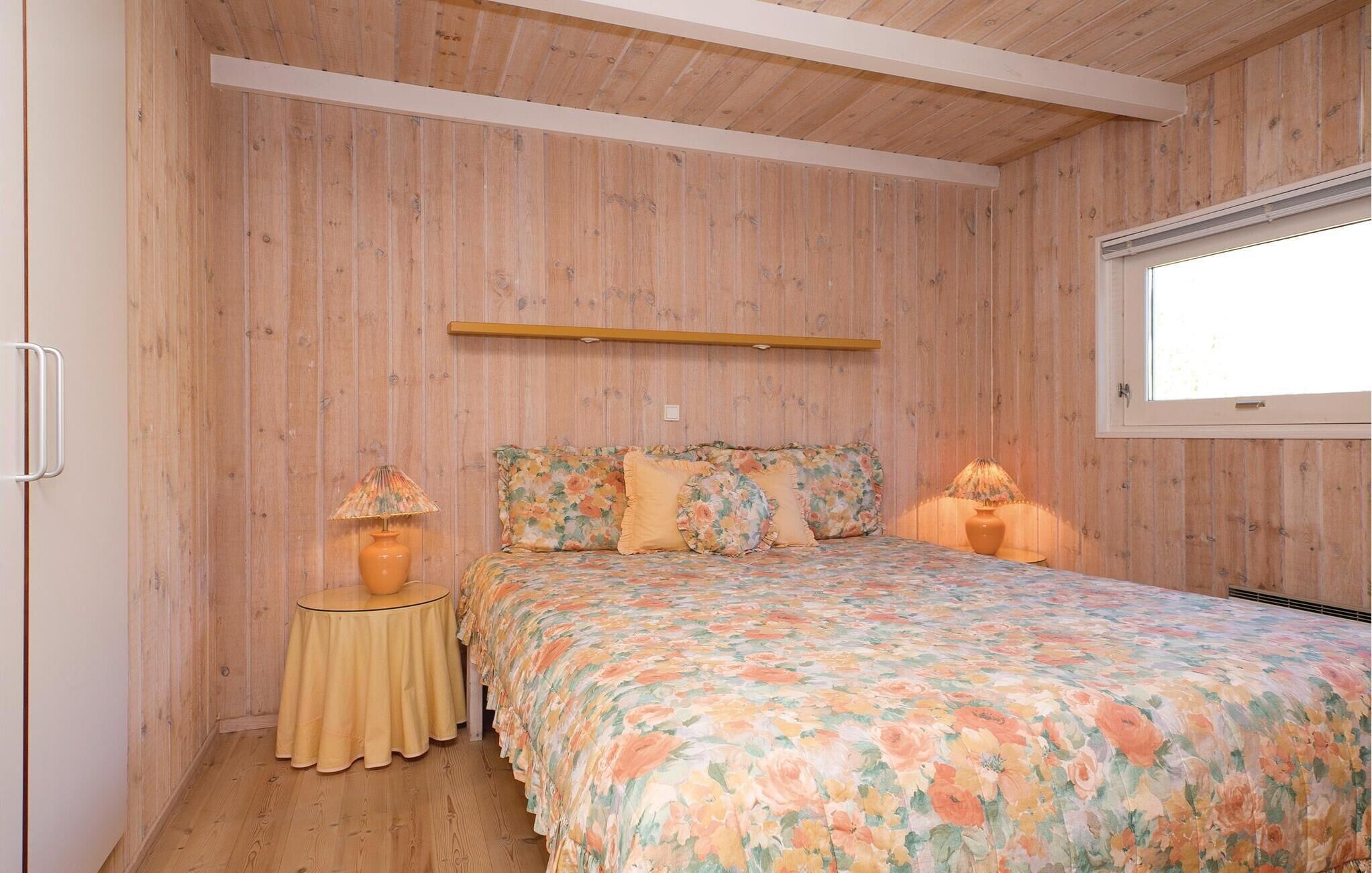A bedroom with a wooden wall panel, a bed with floral bedding, two bedside lamps, and a window.