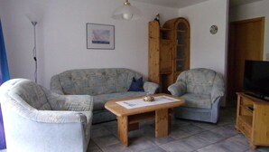 A living room with a sofa, two armchairs, a wooden coffee table, a television on a wooden stand, a ceiling fan, and a framed picture on the wall. - Apartment No. 5 - To the little mermaid (Friedrichskoog)