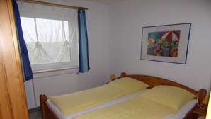 A bedroom with a wooden bed, a window with blue curtains, a radiator, and a framed picture on the wall. - Apartment No. 5 - To the little mermaid (Friedrichskoog)