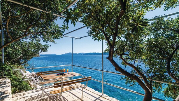 Una terraza con bancos de madera que da a un mar azul y despejado, con vista a montañas distantes.