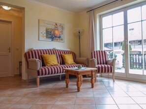 A living room with a striped sofa, a wooden coffee table, and a large window with a view of buildings. - Residence Laguna Whg L-06 .. - L-06 (Kühlungsborn)