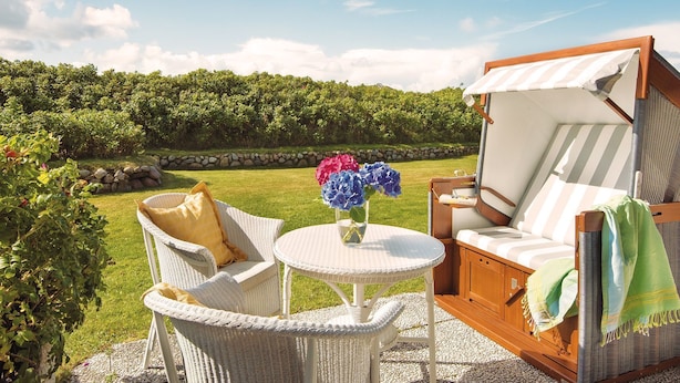 A wicker outdoor seating area with a table and chairs, a cushioned bench, and a view of a vineyard. - Kampen-Kliff 3 from A&M - Kampen-Kliff 3 (Kampen (Sylt))