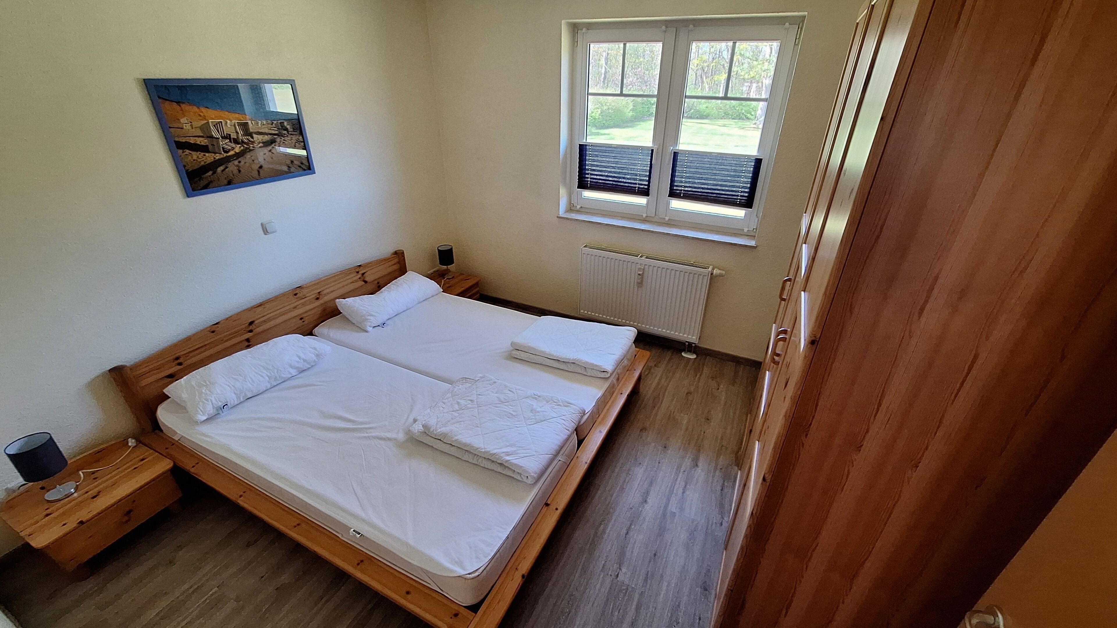 Ein Doppelbett mit weißen Leinen, ein Holz-Kopfteil, ein Nachttisch mit Lampe und ein Fenster mit Jalousien.