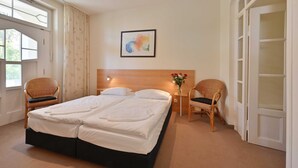 A hotel room with a double bed, two wicker chairs, a nightstand with a vase of flowers, and a framed abstract painting on the wall. - Apartment near the beach 01 Villa Celia 6 pers. - Villa Celia Sellin (Sellin)