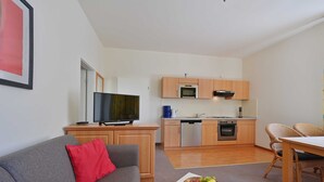 A modern living room with a grey sofa, a red pillow, and a fruit bowl on a small table. A kitchen with wooden cabinets and appliances is visible in the background. - Apartment near the beach 01 Villa Celia 6 pers. - Villa Celia Sellin (Sellin)