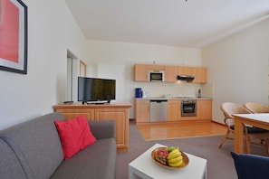 A modern living room with a grey sofa, a red pillow, and a fruit bowl on a small table. A kitchen with wooden cabinets and appliances is visible in the background.
