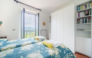 Un dormitorio con un ventanal grande, una cama con ropa de cama floral, un radiador y una estantería llena de libros.