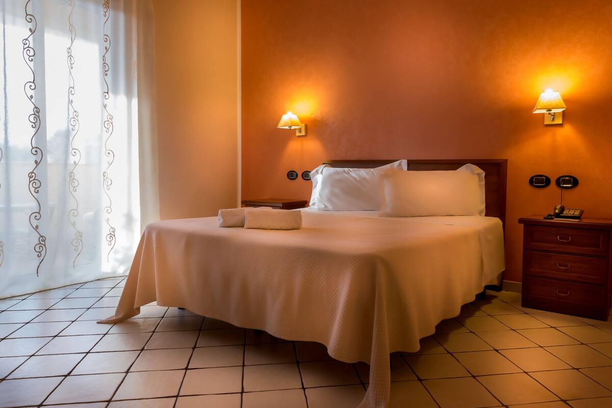 Una cama bien hecha con ropa blanca, dos mesitas de noche con lámparas y una ventana con cortinas.
