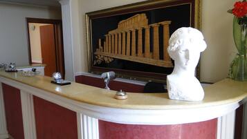 A reception desk with a marble top, a bust sculpture, a framed painting of ancient columns, and a vase with red flowers.