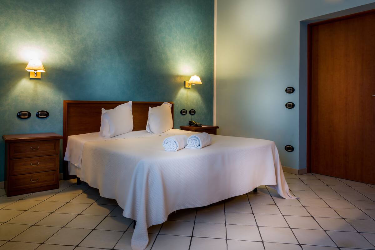 Una cama bien hecha con sábanas blancas y dos toallas enrolladas, una mesita de noche de madera con una lámpara, y una puerta de madera.