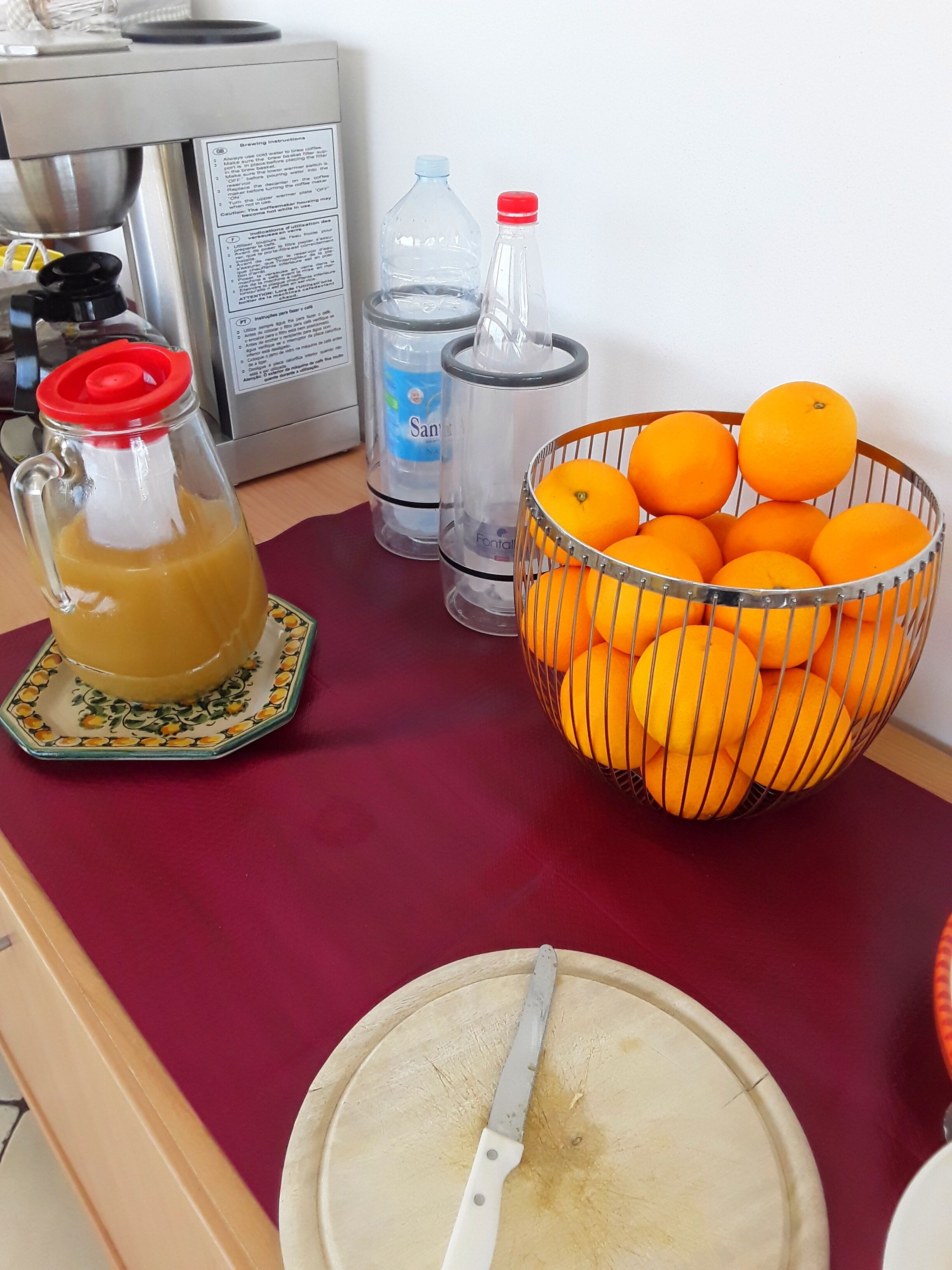 Un comptoir de cuisine avec un panier de fruits contenant des oranges, une carafe de jus et deux bouteilles d’eau.