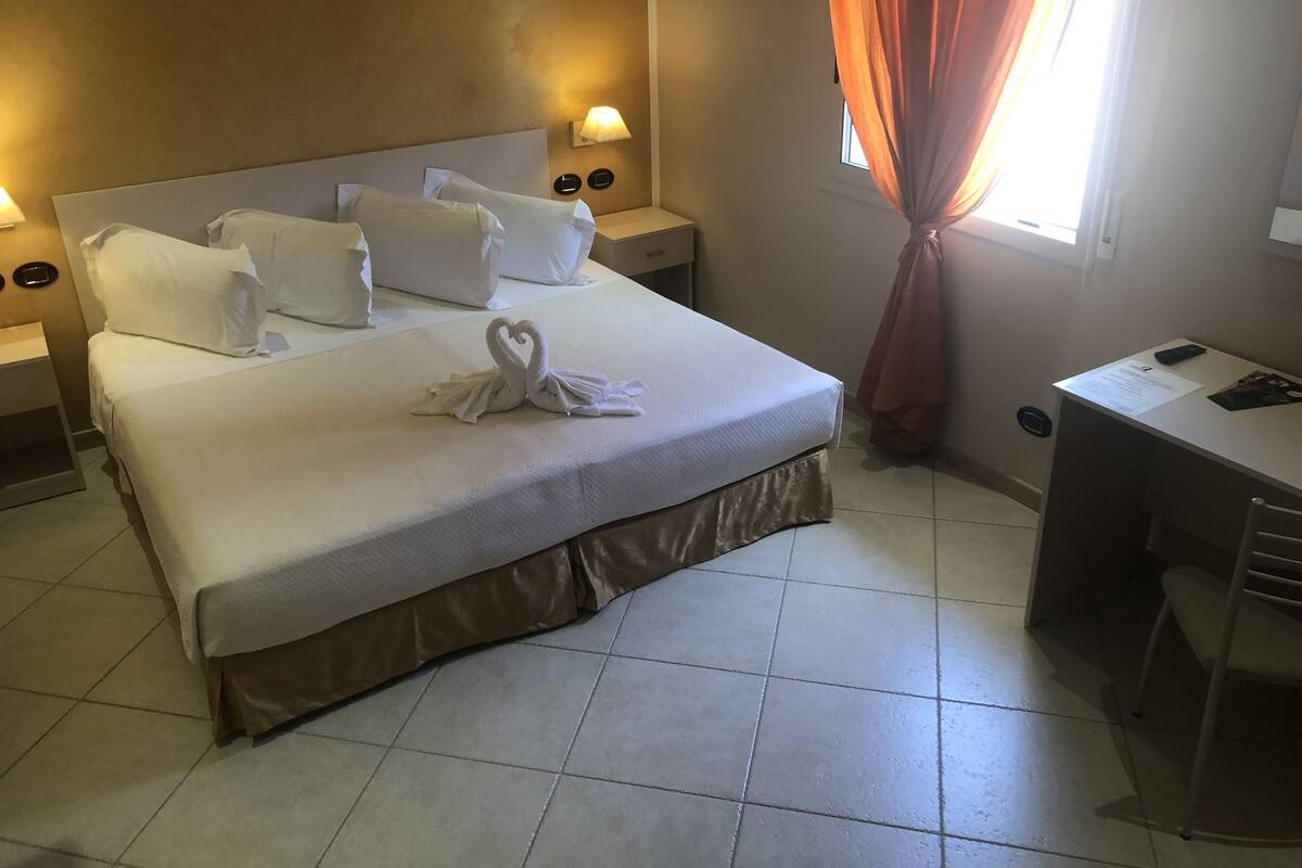 Una cama bien tendida con sábanas blancas y una toalla dispuesta en forma de corazón, un escritorio con una silla y una ventana con cortinas.