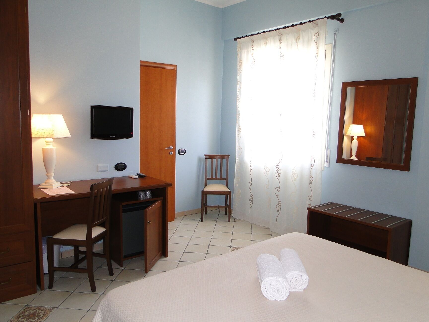 A hotel room with a bed, a desk with a chair, a television, a door, and a window with curtains.