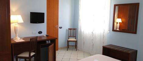A hotel room with a bed, a desk with a chair, a television, a door, and a window with curtains.
