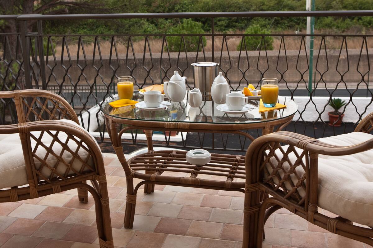 Una mesa de vidrio con sillas de mimbre dispuesta para el desayuno, con una cafetera, tazas, un jarro y jugo de naranja.