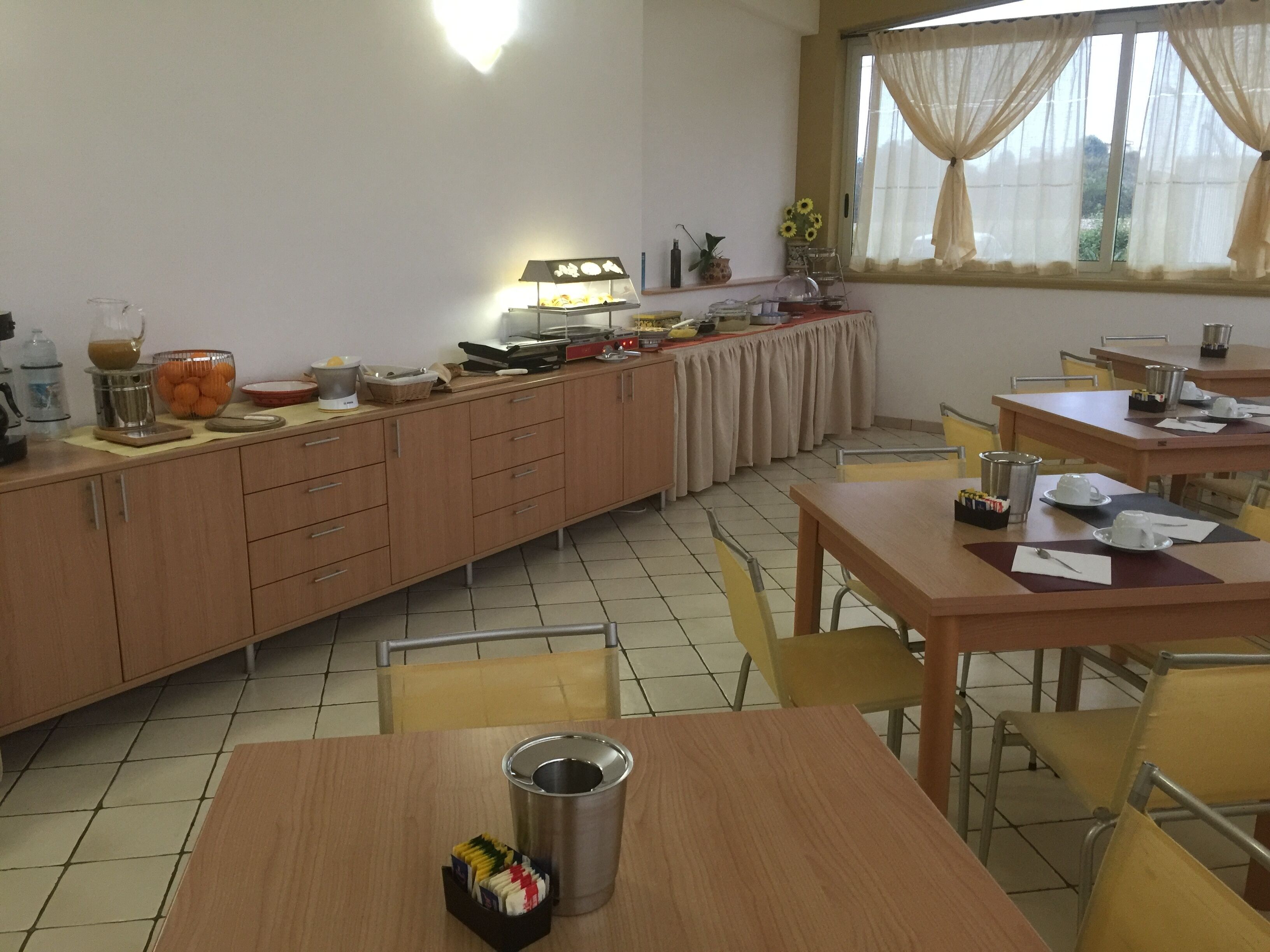 A dining area with wooden tables and chairs, a buffet setup with food items, and a window with curtains.