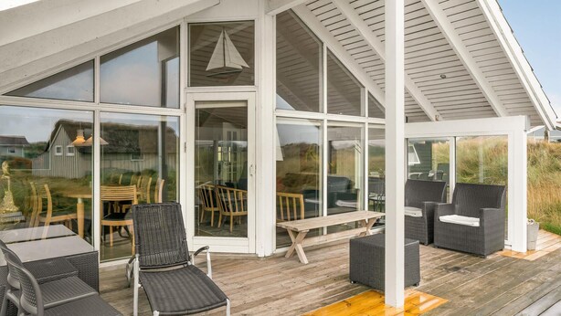 A covered outdoor seating area with wicker furniture and a wooden table, large glass doors leading to an interior space, and a wooden deck. - Beach front home in Fanø with sauna (Fanø)