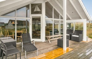 A covered outdoor seating area with wicker furniture and a wooden table, large glass doors leading to an interior space, and a wooden deck. - Beach front home in Fanø with sauna (Fanø)