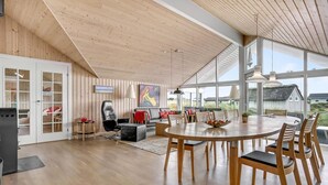 A spacious living room with a large wooden ceiling, a round dining table, and a view of the outdoors through large windows. - Beach front home in Fanø with sauna (Fanø)