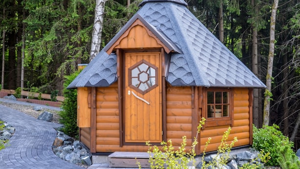 A small wooden cabin with a hexagonal door and a steeply pitched roof, surrounded by a forested area.
