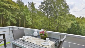 A balcony with a table set for two, a wicker chair, and a potted plant with flowers.