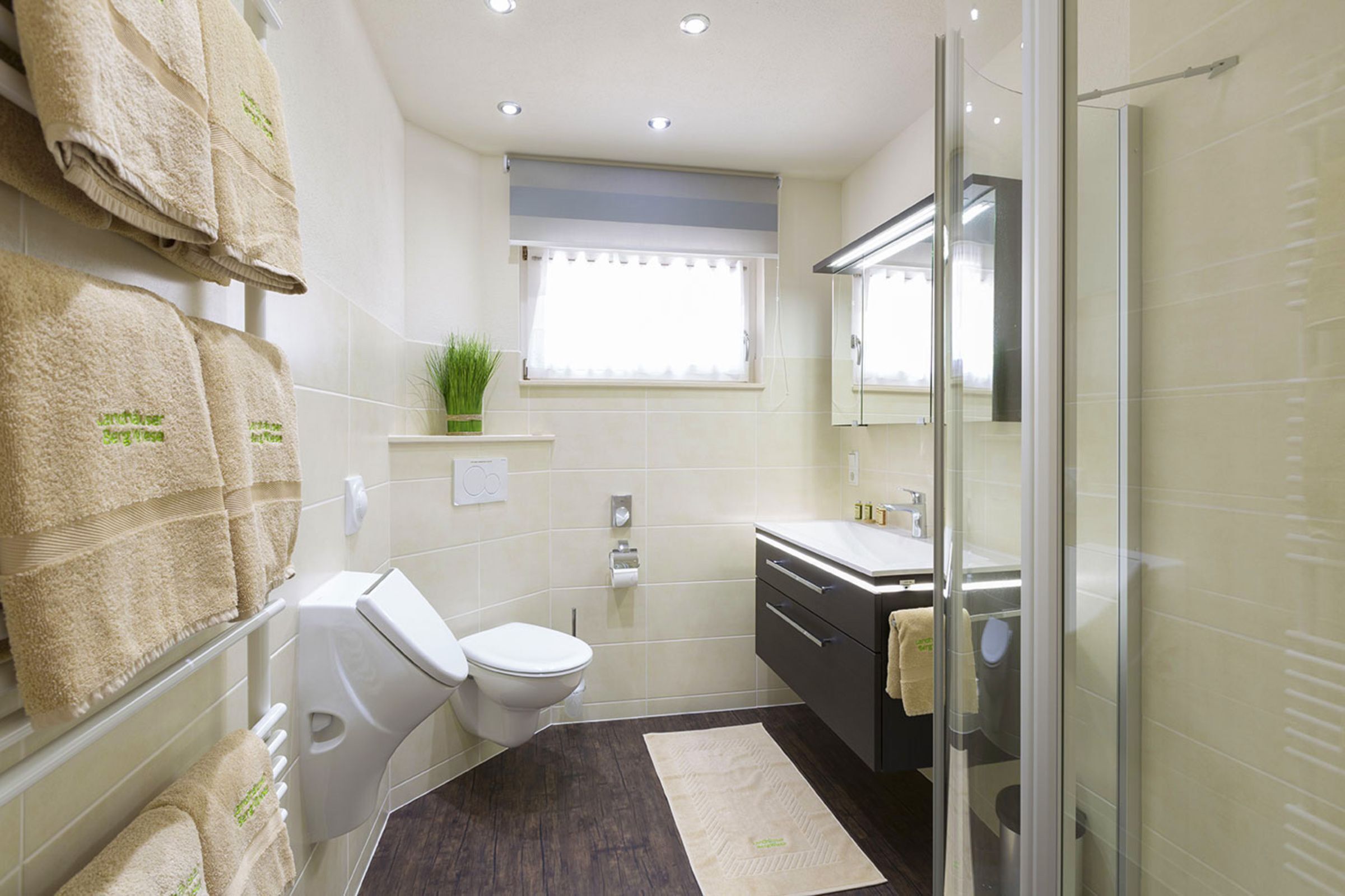 Ein modernes Badezimmer mit Toilette, Waschbecken und Duschbereich. Handtücher hängen an einem Wäscheständer und auf der Fensterbank steht eine kleine Pflanze.