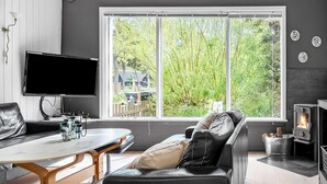 Ett modernt vardagsrum med ett stort fönster, en platt-TV, en soffa, ett matbord och en vedkamin.