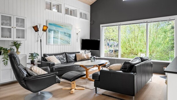 Ett modernt vardagsrum med en svart lädersoffa, ett runt soffbord och en tv som är monterad på väggen. Det finns flera fönster som släpper in naturligt ljus och ger utsikt över grönområden utanför.