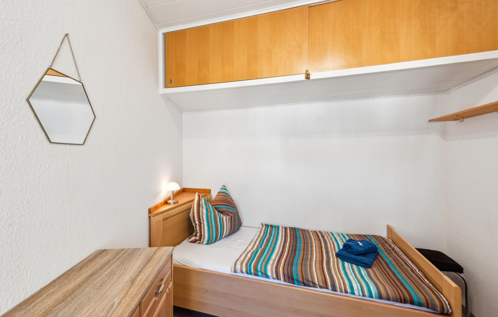 Ein gemütliches Schlafzimmer mit einem Holzbett, gestreifter Bettwäsche, einem Nachttisch mit Lampe, einem Hänge-Spiegel und einer kleinen Holzkommode.