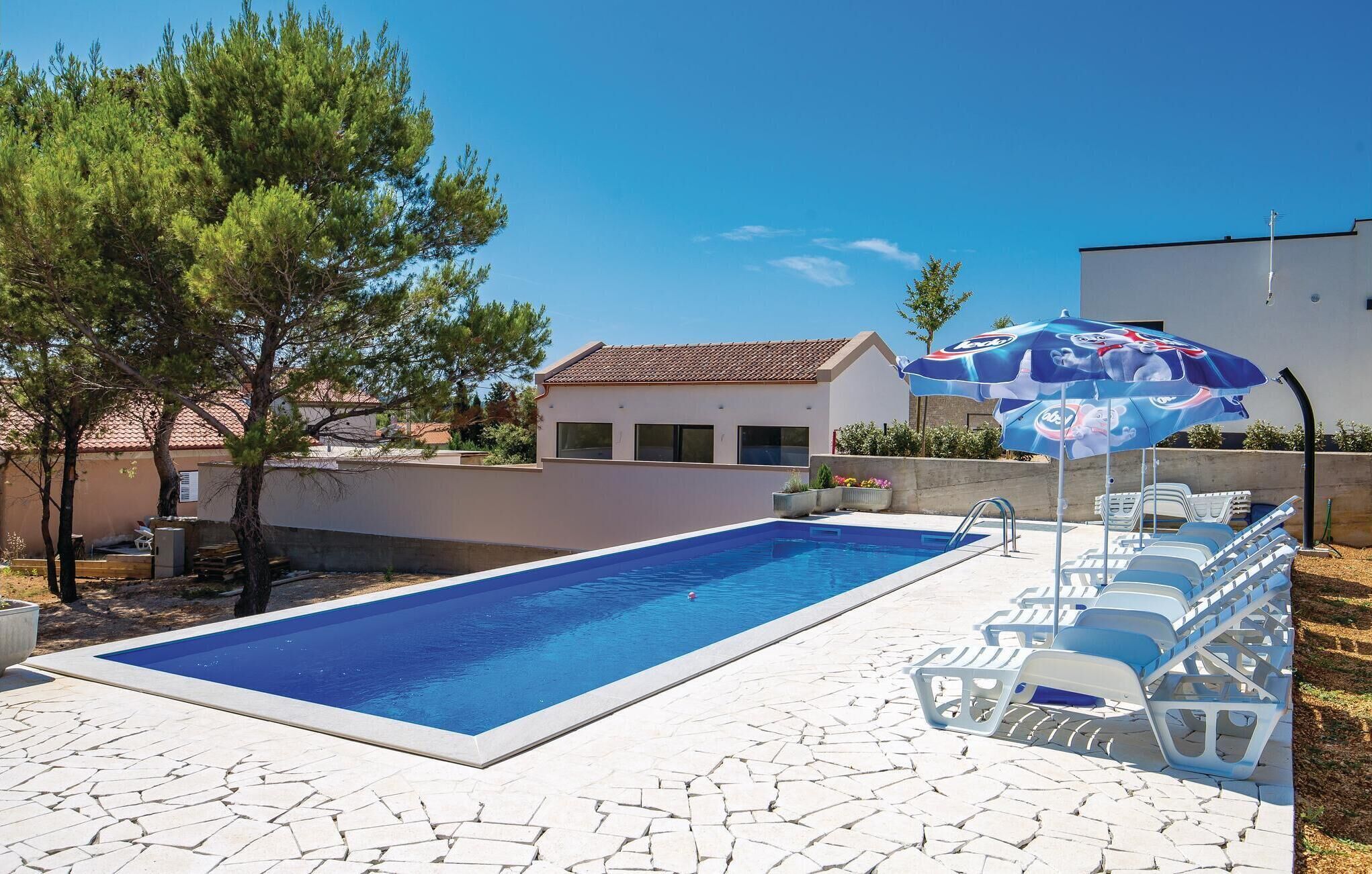 A rectangular swimming pool with a tiled deck, surrounded by lounge chairs and an umbrella.