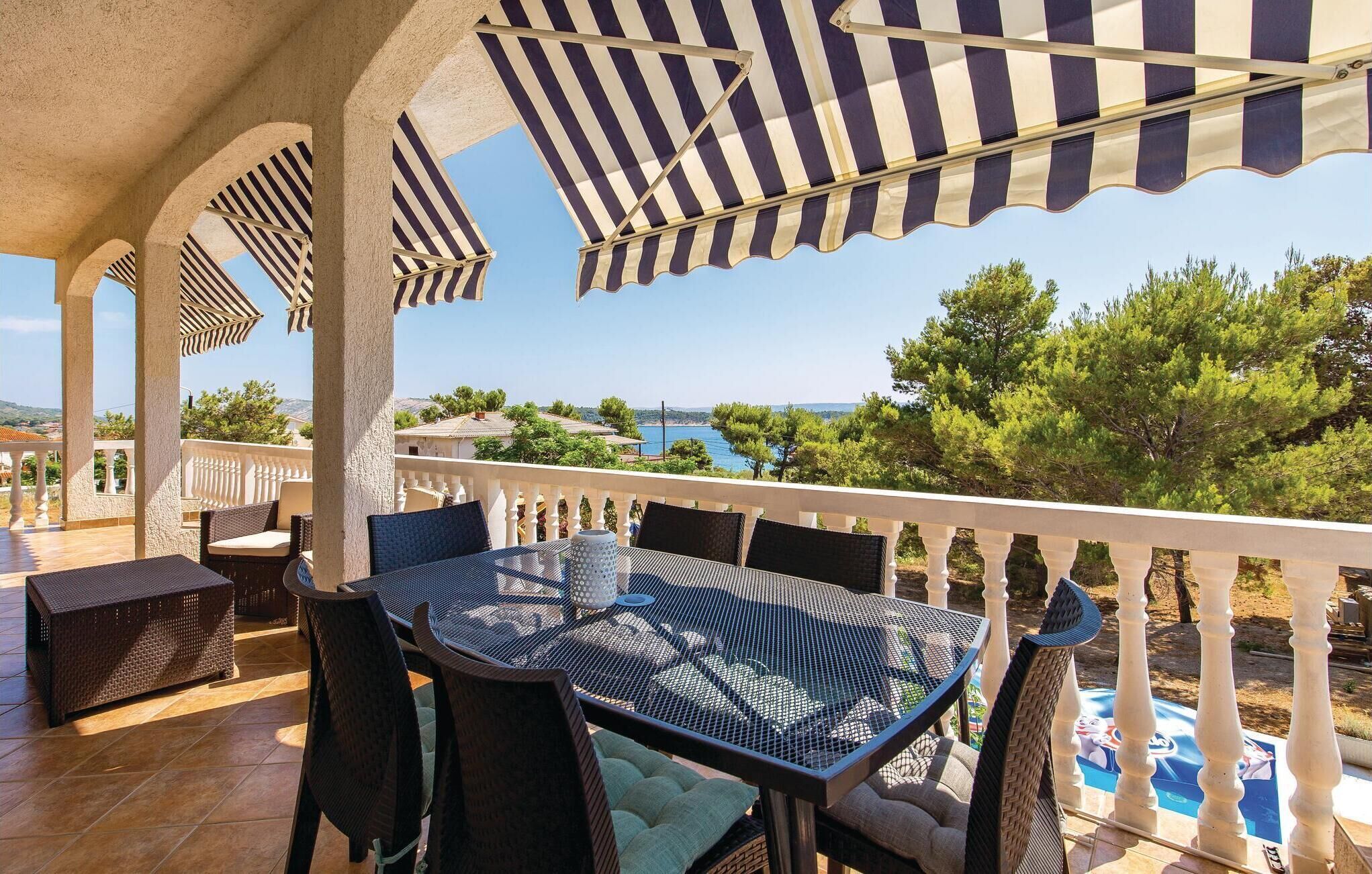 A covered outdoor seating area with a table and chairs, overlooking a scenic view of the sea and greenery.
