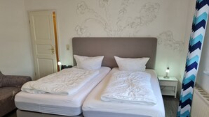 Een tweepersoonsbed met witte beddengoed, een bruin hoofdeinde, een nachtkastje met een lamp, een stoel, en een muur met een bloemenmural.