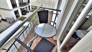 Een balkon met een ronde glazen tafel en twee rieten stoelen, met uitzicht op een binnenplaats met een gebouw op de achtergrond.