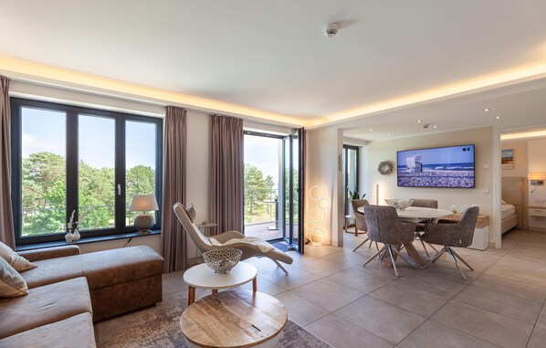 A modern living room with a large window, a dining area, and a flat-screen TV mounted on the wall.