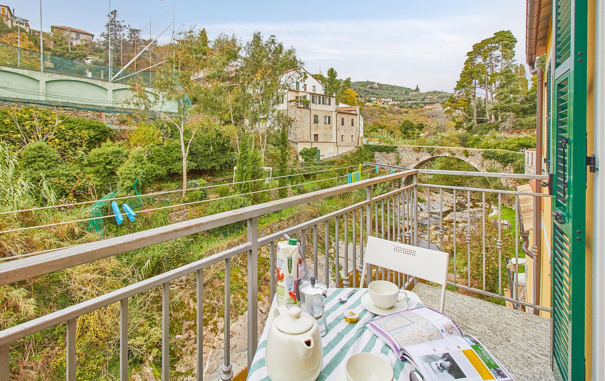 Un balcón con una mesa lista para el té, con vistas a un puente de piedra y abundante vegetación.