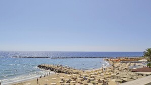 A beach with numerous umbrellas and sun loungers, a rocky breakwater, and a clear blue sky.