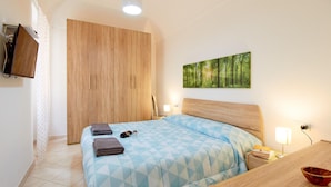 A bedroom with a bed, a wooden wardrobe, a TV mounted on the wall, and a painting of a forest.