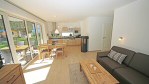 A modern living room with a sofa, coffee table, and dining area with chairs. A kitchen is visible in the background with cabinets and appliances. - Vacation home Igelbau Whg. 01 with terrace - MR: Holiday home Igelbau with 2 (Baabe)