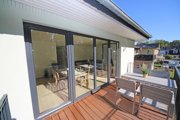 Een modern huis met een houten terras, grote schuifpuien die leiden naar een buiten eethoek met een tafel en stoelen.